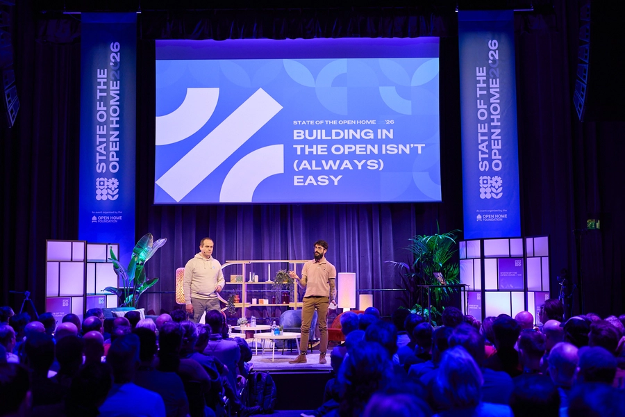Franck, Home Assistant Lead, and JLo, Product and Design Lead, standing onstage presenting to a large audience, with a screen behind them reading Building in the open isn't (always) easy, and a softly lit set with plants and shelves.