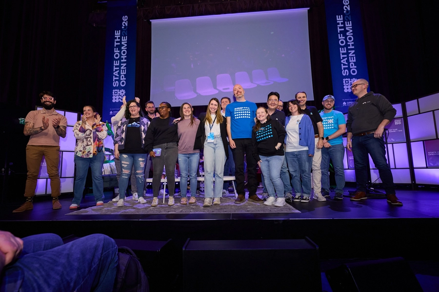 A group of speakers and organizers standing together onstage, smiling and applauding in front of a large screen and event banners, marking the close of the event.